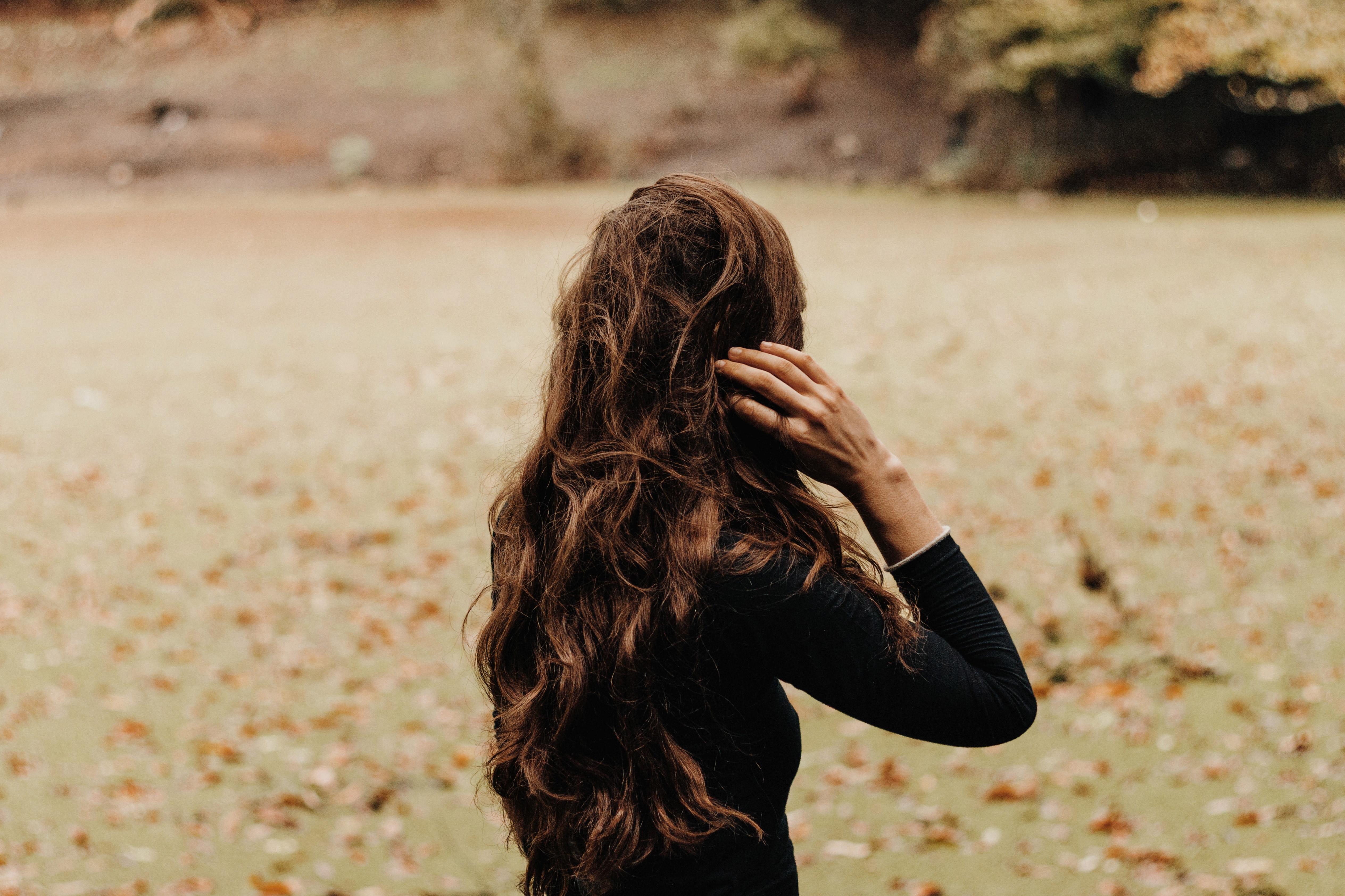 Photo of the back of a brunette woman with long hair wearing a black sweater standing on a green lawn full of fall leaves
