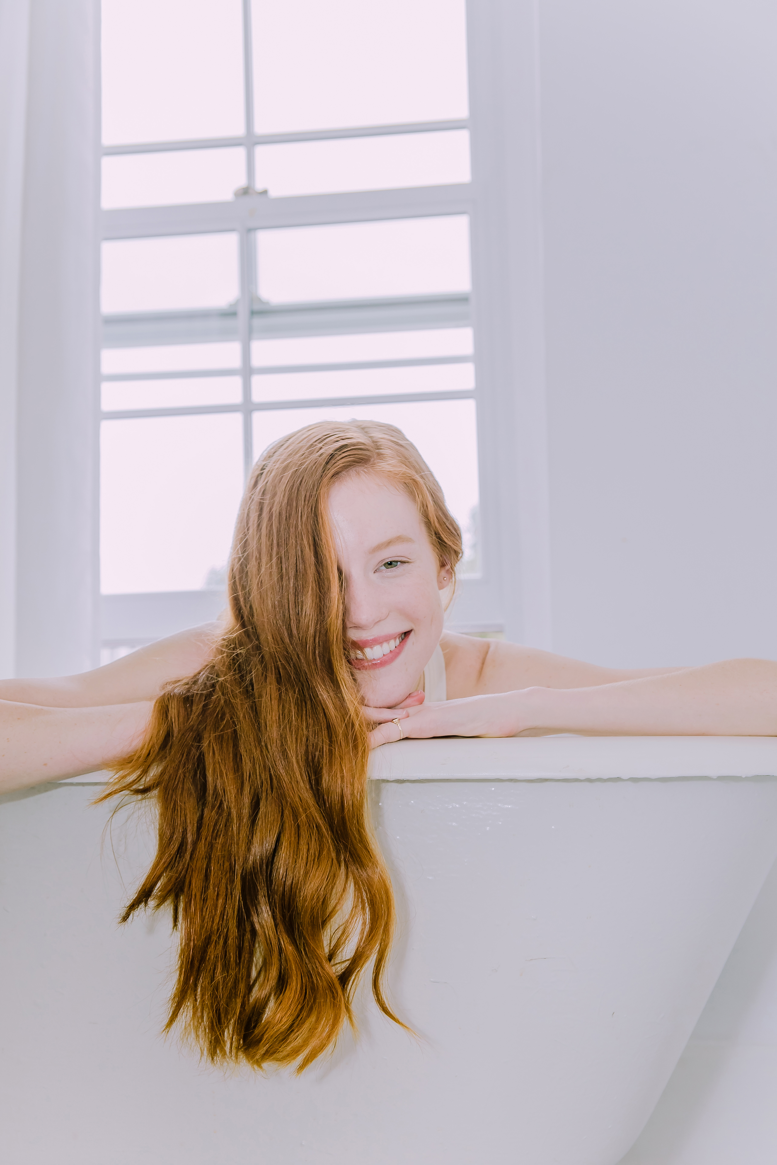 ginger haired woman in an all white bathtub smiling with her hair in her face