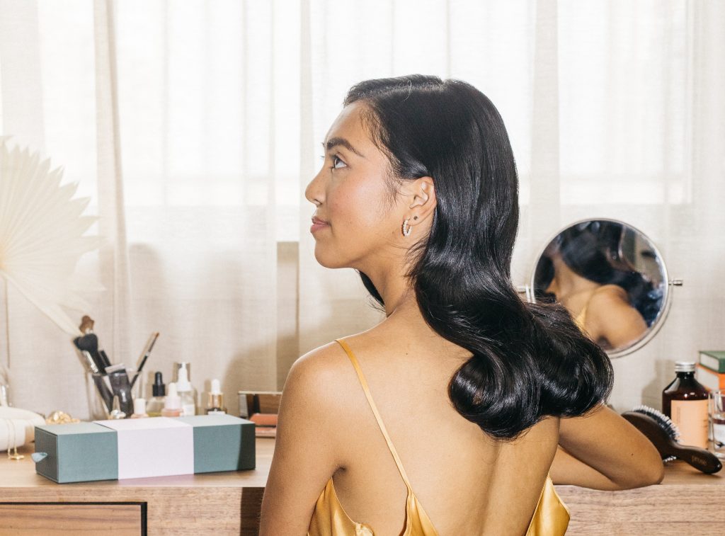 a woman with dark, sleek hair sits at her vanity in a yellow, silk slip dress