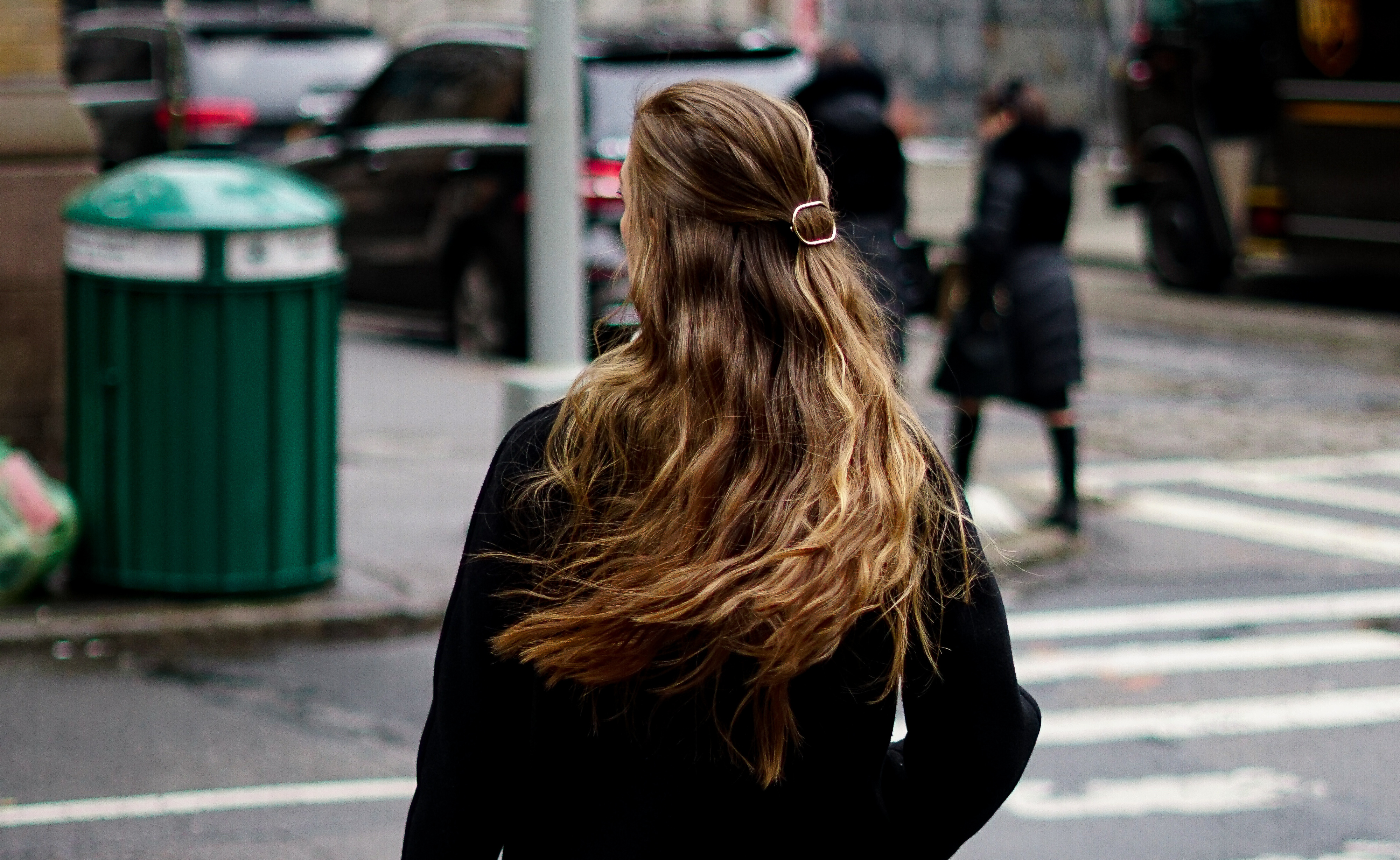 a woman with very long honey blonde hair stands in a busy NYC street wearing a black coat