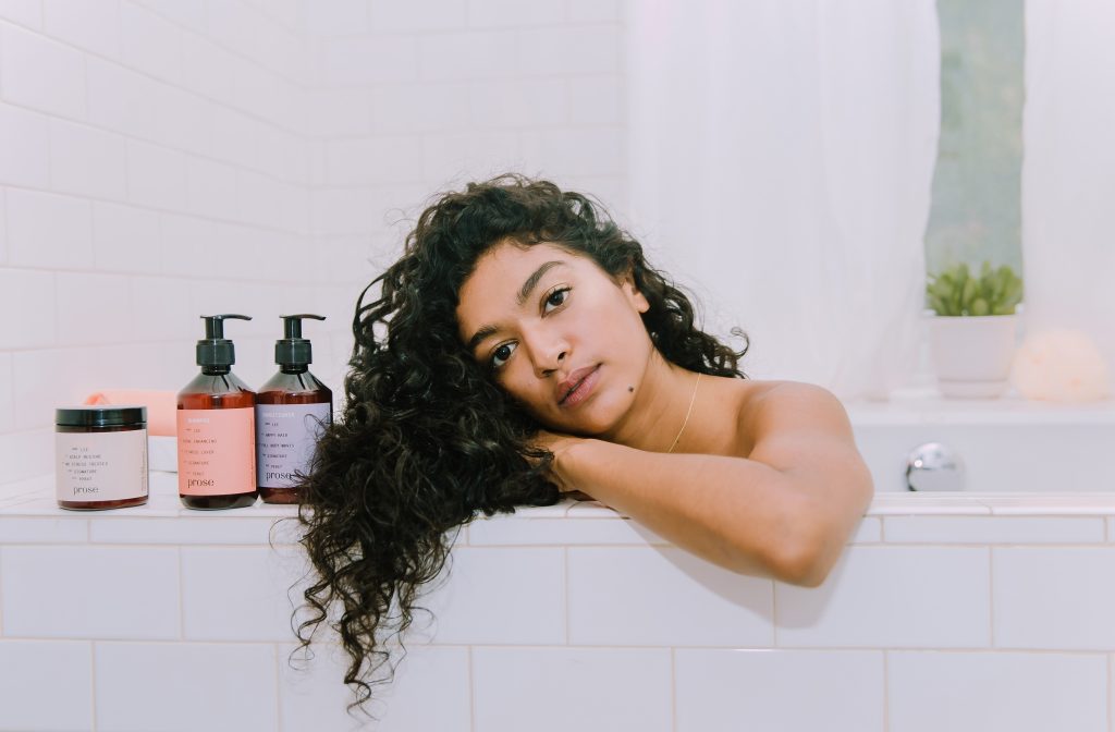 Woman with brown, curly hair in the bathtub with her Prose products