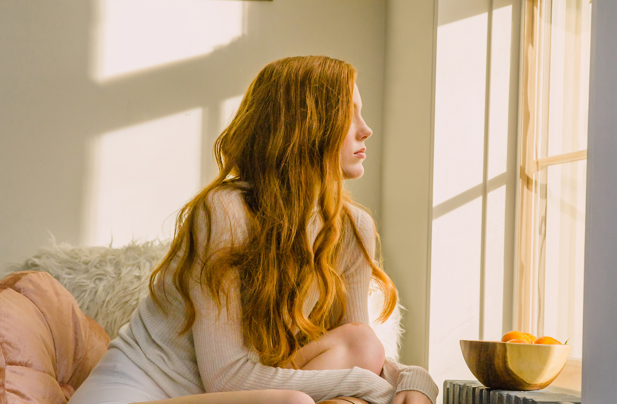 Prose model with long, red, wavy hair looking out a window with soft sunlight flooding in