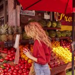 Prose model with long, blonde hair shopping in a red top at a fruit stand