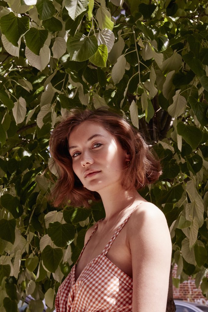 Prose model with short, brown hair posing in front of leafy tree in a red and white dress
