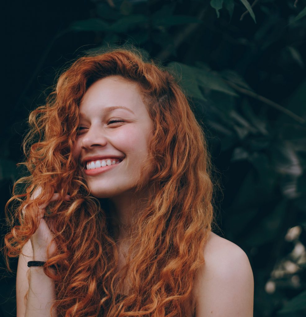 Girl with curly, red hair smiling against greenery