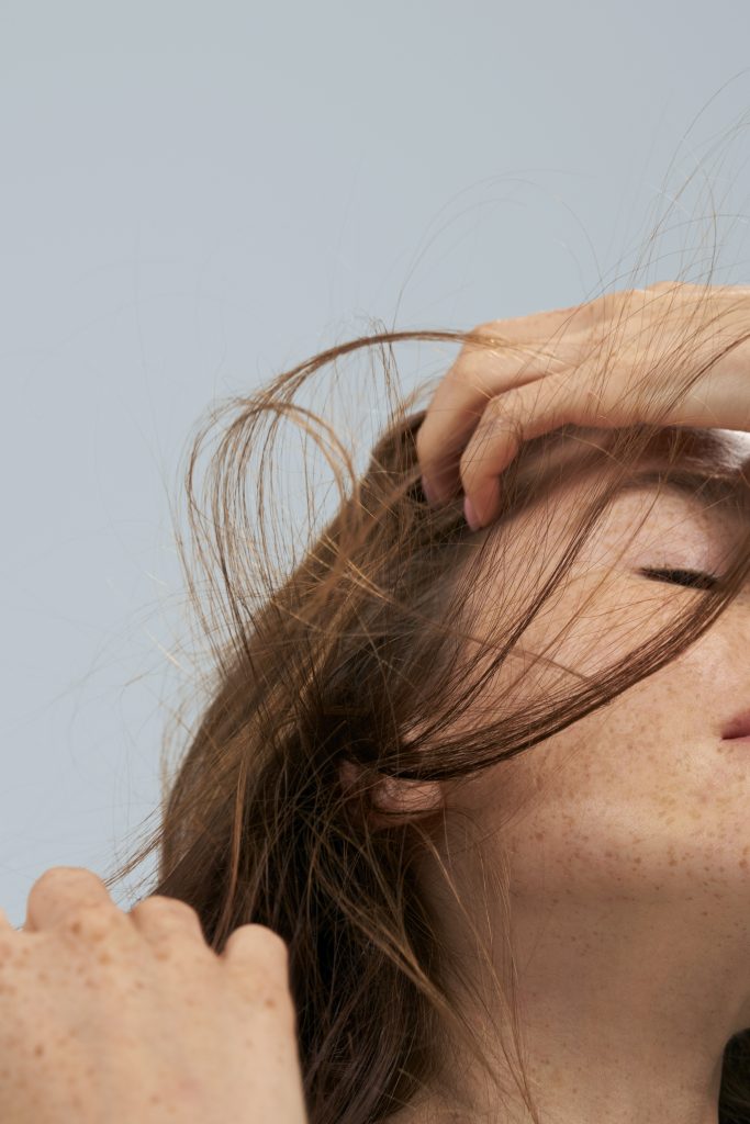 woman with auburn hair and a freckled complexion runs her fingers through her hair as a gust of wind blows her strands