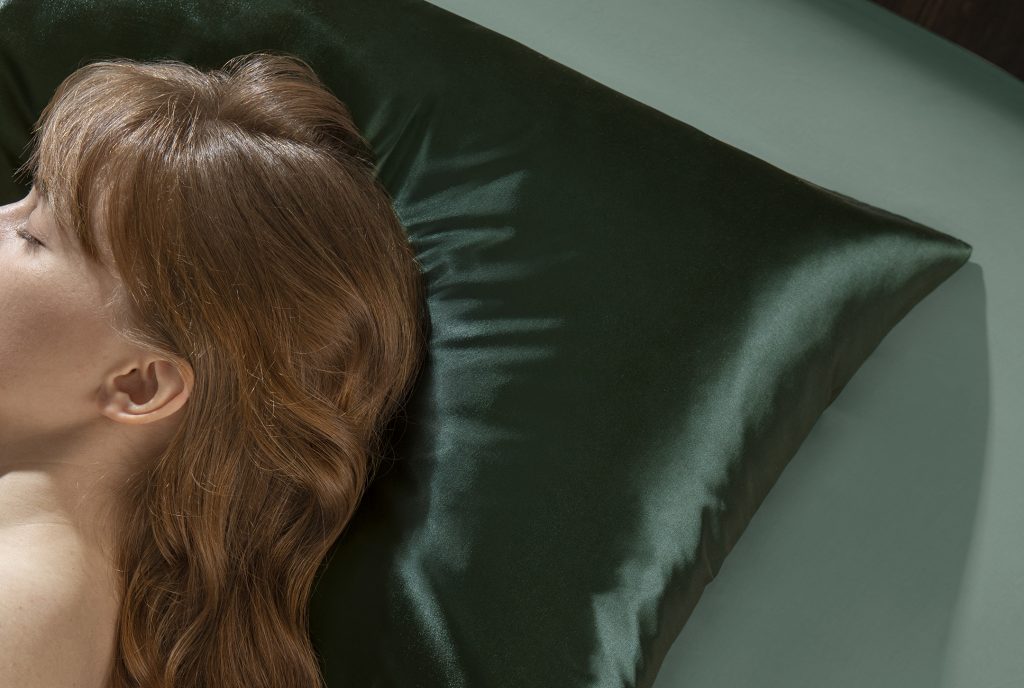 woman with wavy brown hair and bangs sleeping on a dark green silk pillowcase