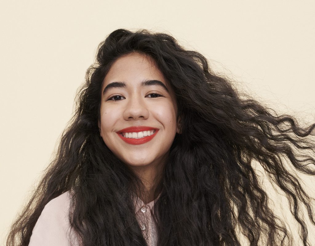 girl with long, black wavy hair smiles at the camera while wearing red lipstick