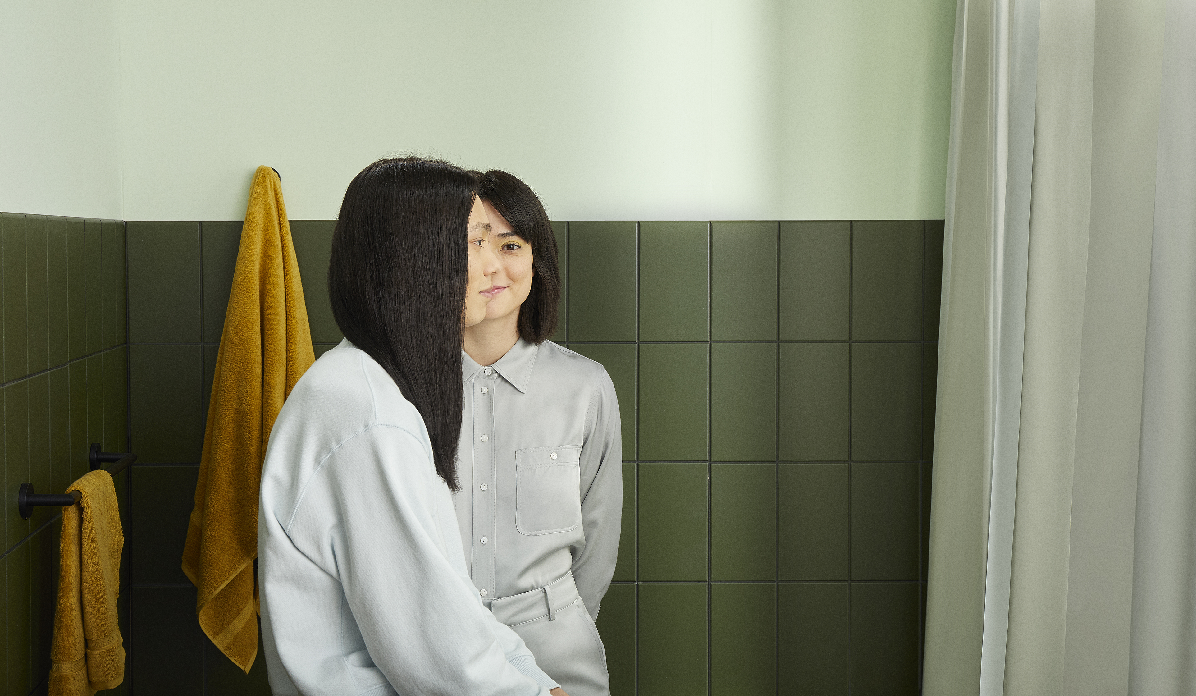 man with long black hair and woman with short black hair stand in a green, tile bathroom together
