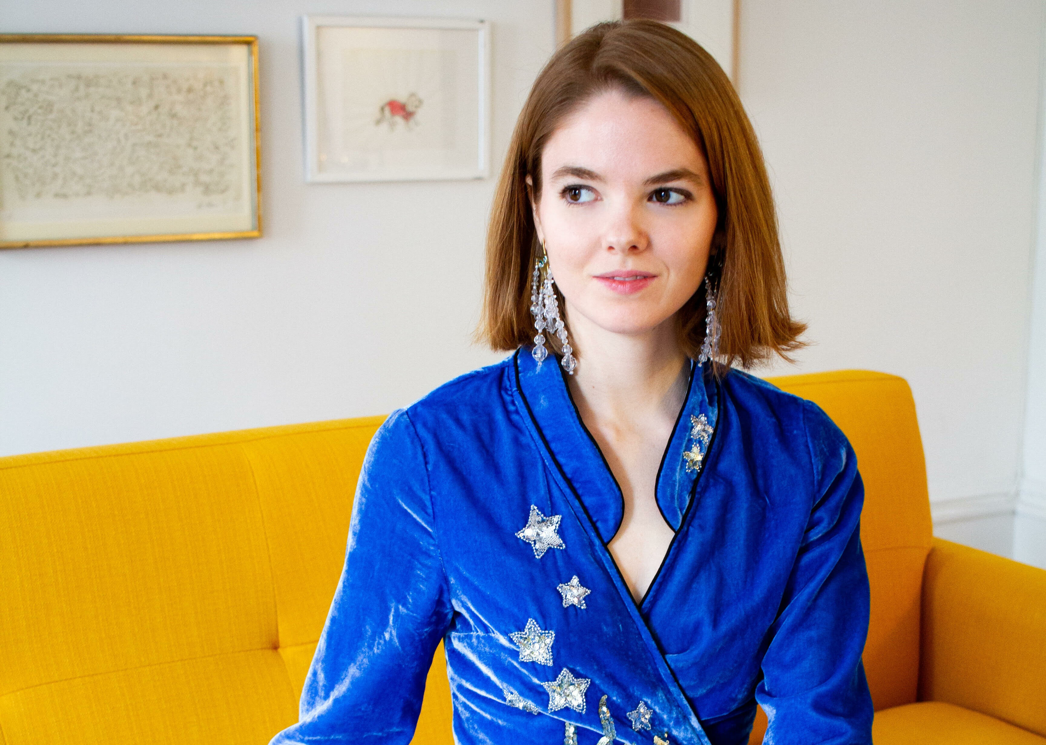 alice bell sits on a yellow couch wearing a blue dress