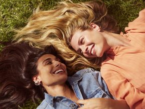 Prose models with long brown and blonde hair laugh while laying in grass