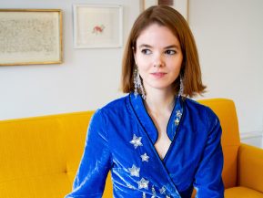 alice bell sits on a yellow couch wearing a blue dress