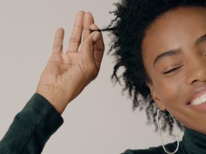 woman with dark coily hair pulls at one of her hair strands while smiling big