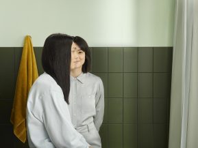 man with long black hair and woman with short black hair stand in a green, tile bathroom together