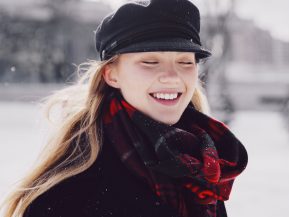 A blonde woman is smiling wearing a black hat, black coat, and red plaid scarf
