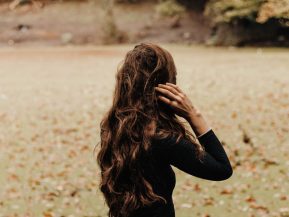 Photo of the back of a brunette woman with long hair wearing a black sweater standing on a green lawn full of fall leaves