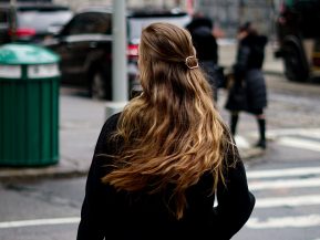 a woman with very long honey blonde hair stands in a busy NYC street wearing a black coat